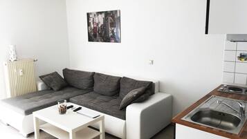 A small, modern apartment with a sofa, a white table, a kitchen area with a sink, and a radiator.