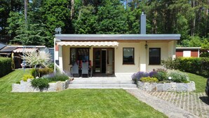 A small, single-story building with a covered porch, a stone pathway leading to the entrance, and a well-maintained garden with various plants. - "Waterkieker" Vacation Home on Lake Jabelschen with a Dock & Outdoor Sauna (Jabel)