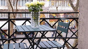 En balkong med ett bord och stolar, en vas med blommor och en utsikt över byggnader i bakgrunden.