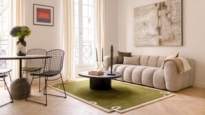 A living room with a round coffee table, two wireframe chairs, a sofa, a green rug, and a painting on the wall. - Harmony Collection I 2BR Suite 4P AC Opera Grevin (Paris)