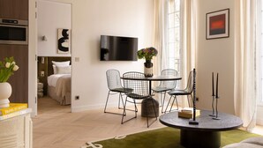 A modern living room with a dining area, a flat-screen TV mounted on the wall, and a large window with curtains. - Harmony Collection I 2BR Suite 4P AC Opera Grevin (Paris)