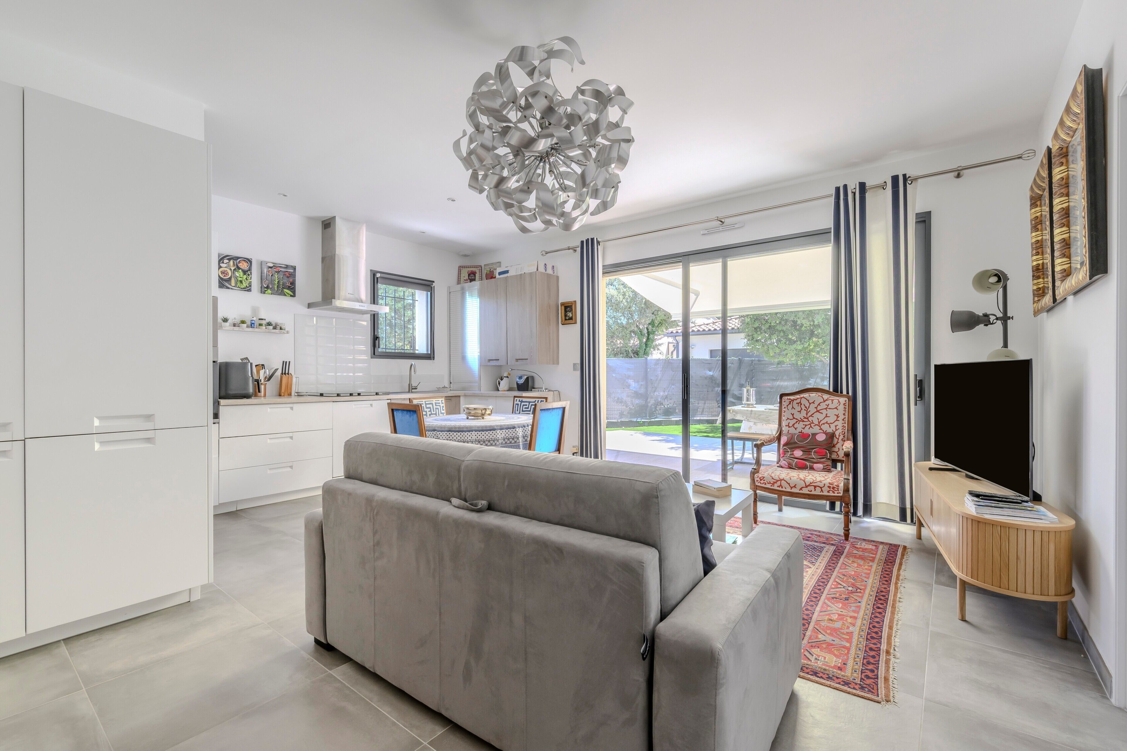 Een moderne keuken met witte kastjes, een zithoek met tafel en stoelen, een grijze bank en een groot raam met uitzicht op de omgeving.