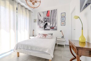 Een slaapkamer met een bed, nachtkastje, lamp, vaas en een muur met ingelijste foto’s.