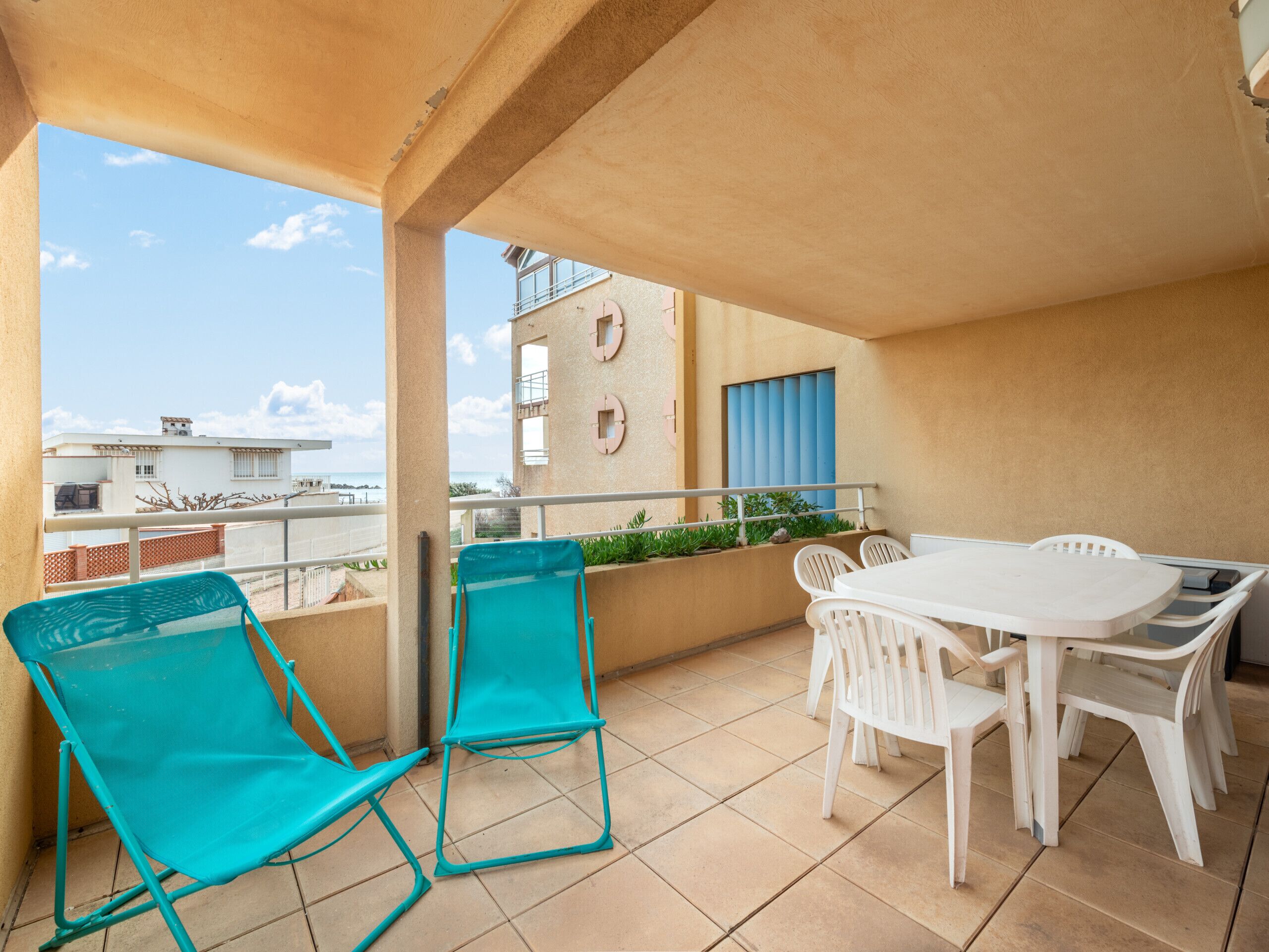 Un balcon avec une table et des chaises blanches, deux chaises turquoise et une vue sur les bâtiments et la mer.