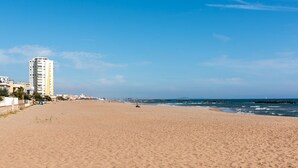 Ein weiter Sandstrand mit einem hohen Gebäude links, blauem Himmel und ruhigen Wellen.