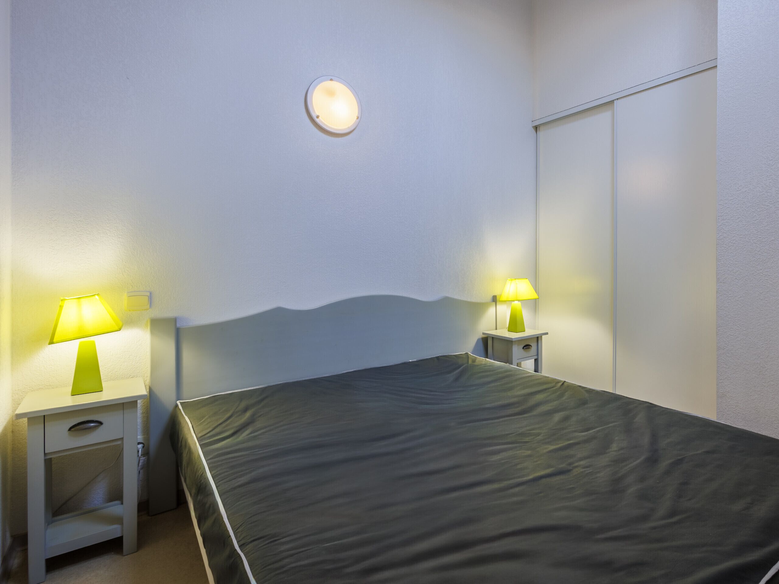 Une chambre à coucher comprenant un lit, deux tables de chevet avec des lampes, un luminaire fixé au mur et une armoire.
