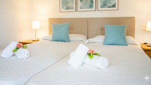 Et hotelværelse med to senge, hver med et hvidt sengetæppe, puder og håndklæder rullet sammen med blomster. Der er to sengeborde med lamper og tre billeder hængende på væggen.