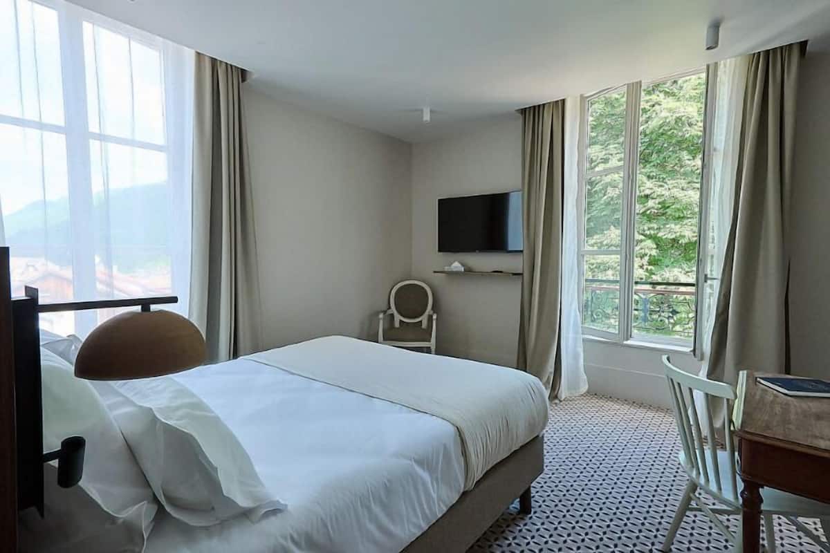 A hotel room with a bed, a desk, a chair, a TV, and large windows.