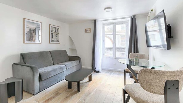 A modern living room with a grey sofa, a glass coffee table, and a TV mounted on the wall. - Studio de charme au cœur du Sentier (Paris)