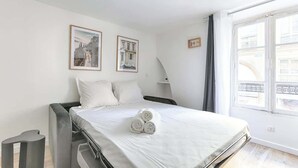 A modern hotel room with a bed, two framed pictures on the wall, a bedside lamp, and a window with a view of buildings. - Studio de charme au cœur du Sentier (Paris)
