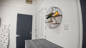 A hallway with a dark door, a white wall with a circular shelf holding decorative items, and a wooden bench. - Charming Studio in Harborne (Birmingham)