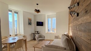 A modern living room with a wooden floor, a sofa, a coffee table, a dining table with chairs, a sewing machine, and a television on the wall.