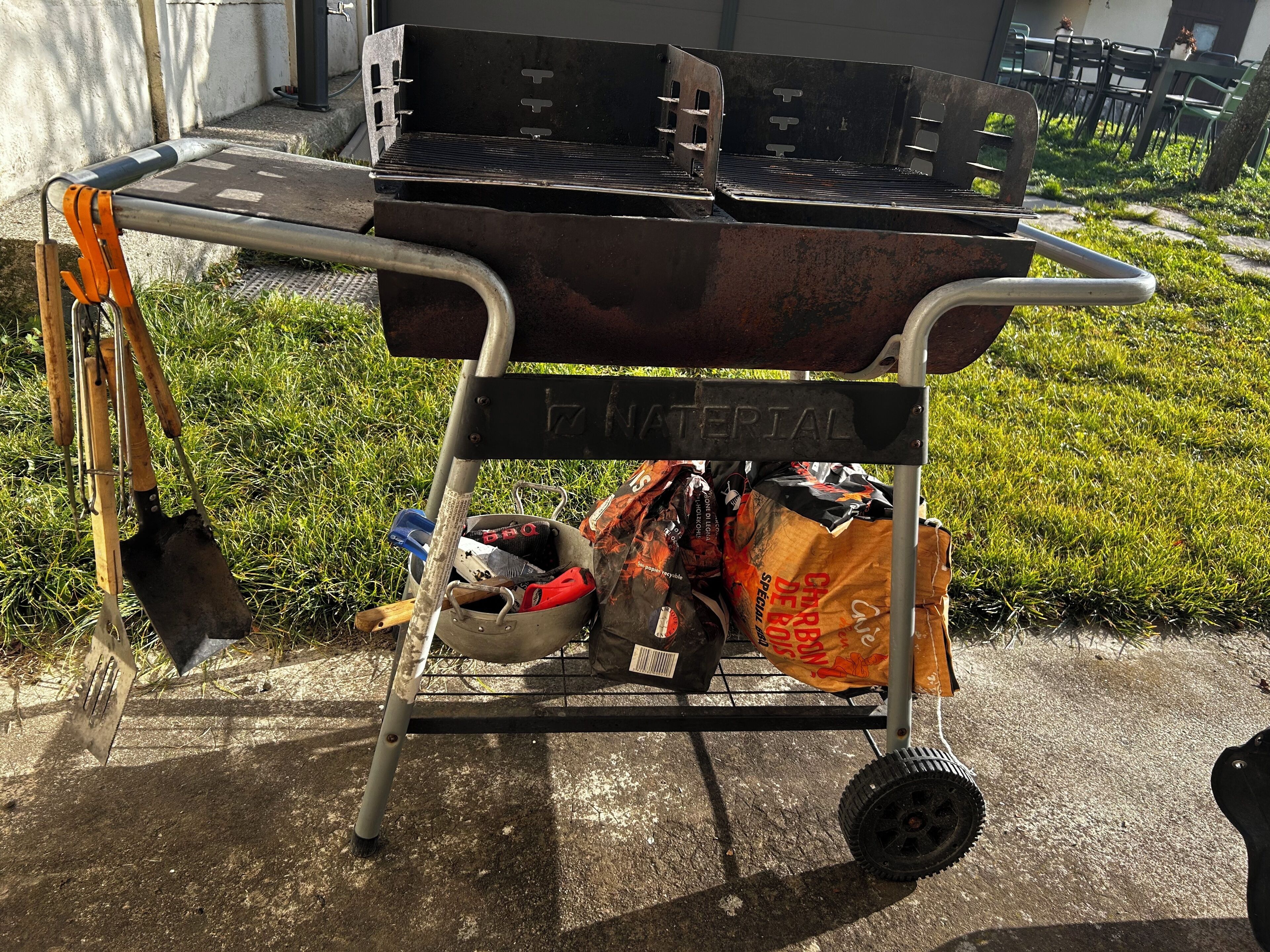 A portable grill cart with a metal frame, labeled "NATURAL," containing a shovel, a pot, and bags of charcoal.
