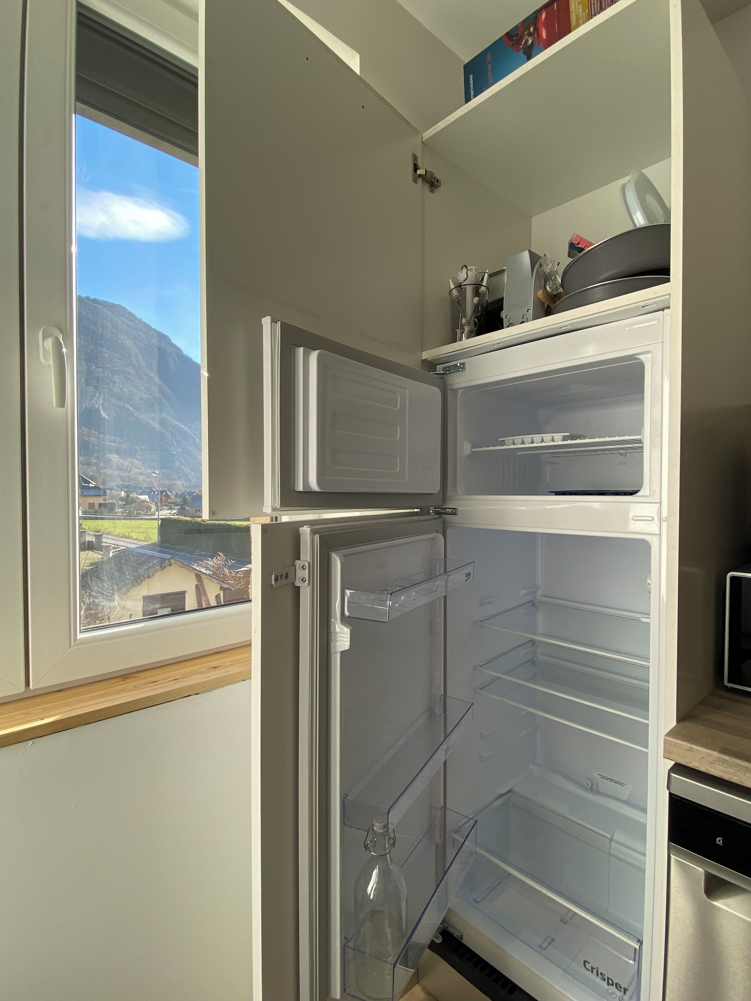 A kitchen with a built-in refrigerator, a window with a view of a mountain, and a shelf with kitchen utensils.