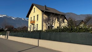 A two-story house with a balcony, surrounded by a low wall and a hedge.
