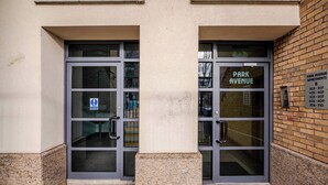 En byggnadsentré med två glasdörrar, en skylt som visar ”Park Avenue Apartments” med lägenhetsnummer, och en liten skylt med telefonnummer till kontaktpersoner i nödsituationer.