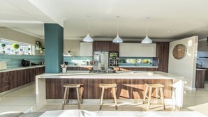 A modern kitchen with a central island, wooden bar stools, pendant lighting, and a large clock on the wall.