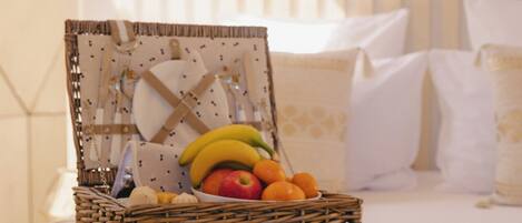 A wicker basket with fruits and two patterned mugs on a bed with white bedding and pillows.