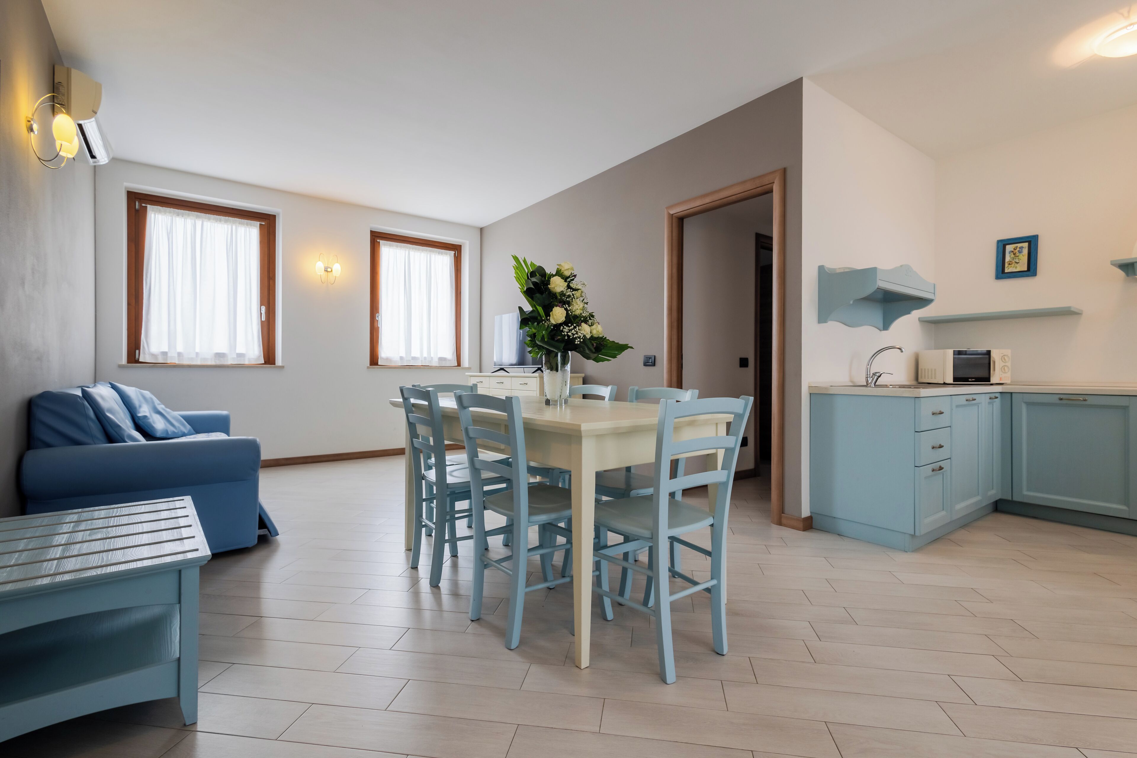 A modern living room with a blue sofa, a dining table with chairs, and a kitchen area with light blue cabinets.