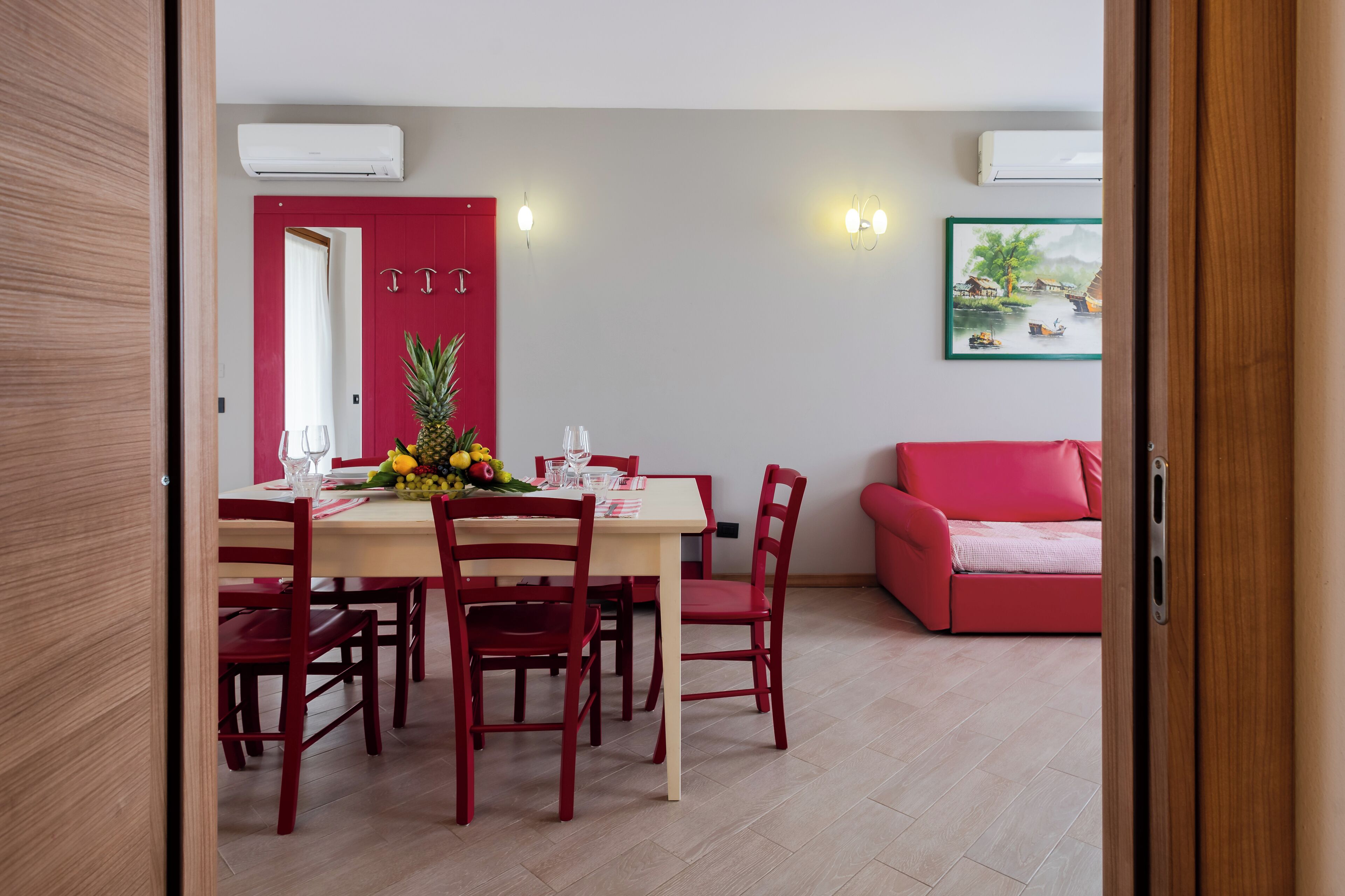 A dining area with a table set for four, surrounded by red chairs, a red sofa, and a painting on the wall.