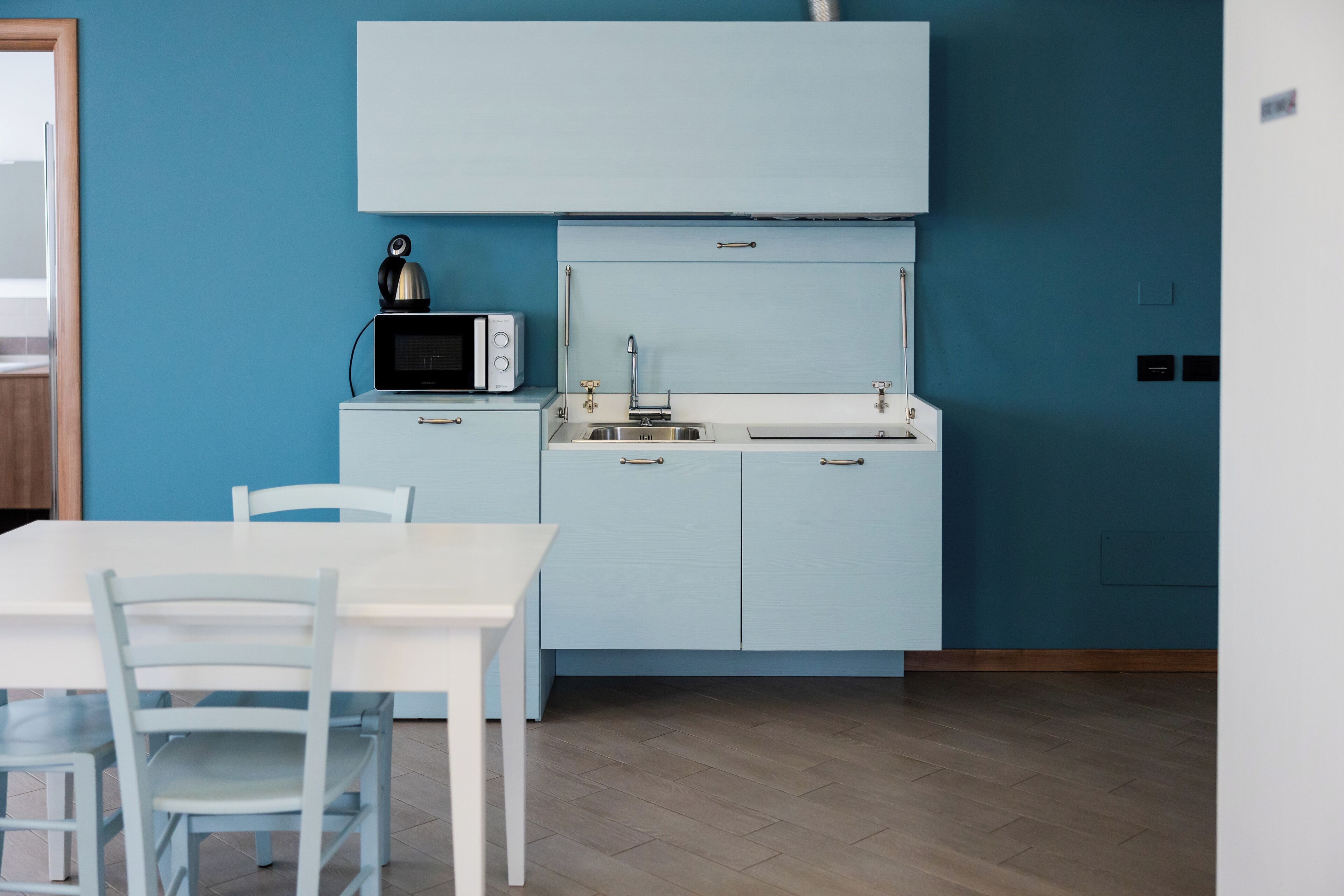 A compact kitchen with a microwave, sink, and cabinets, set against a teal wall.