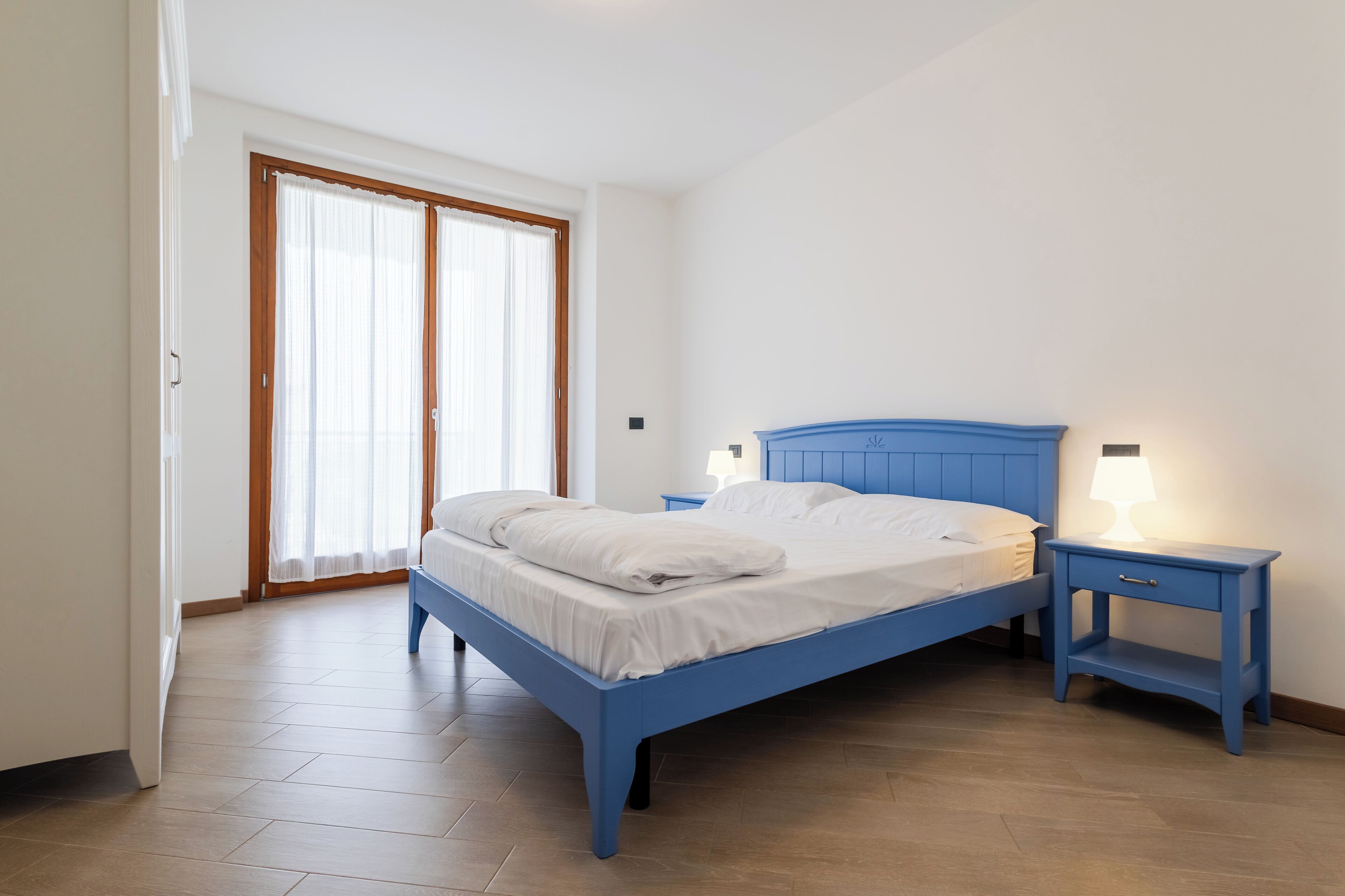 A bedroom with a blue bed, two bedside tables with lamps, a large window with curtains, and a wooden floor.