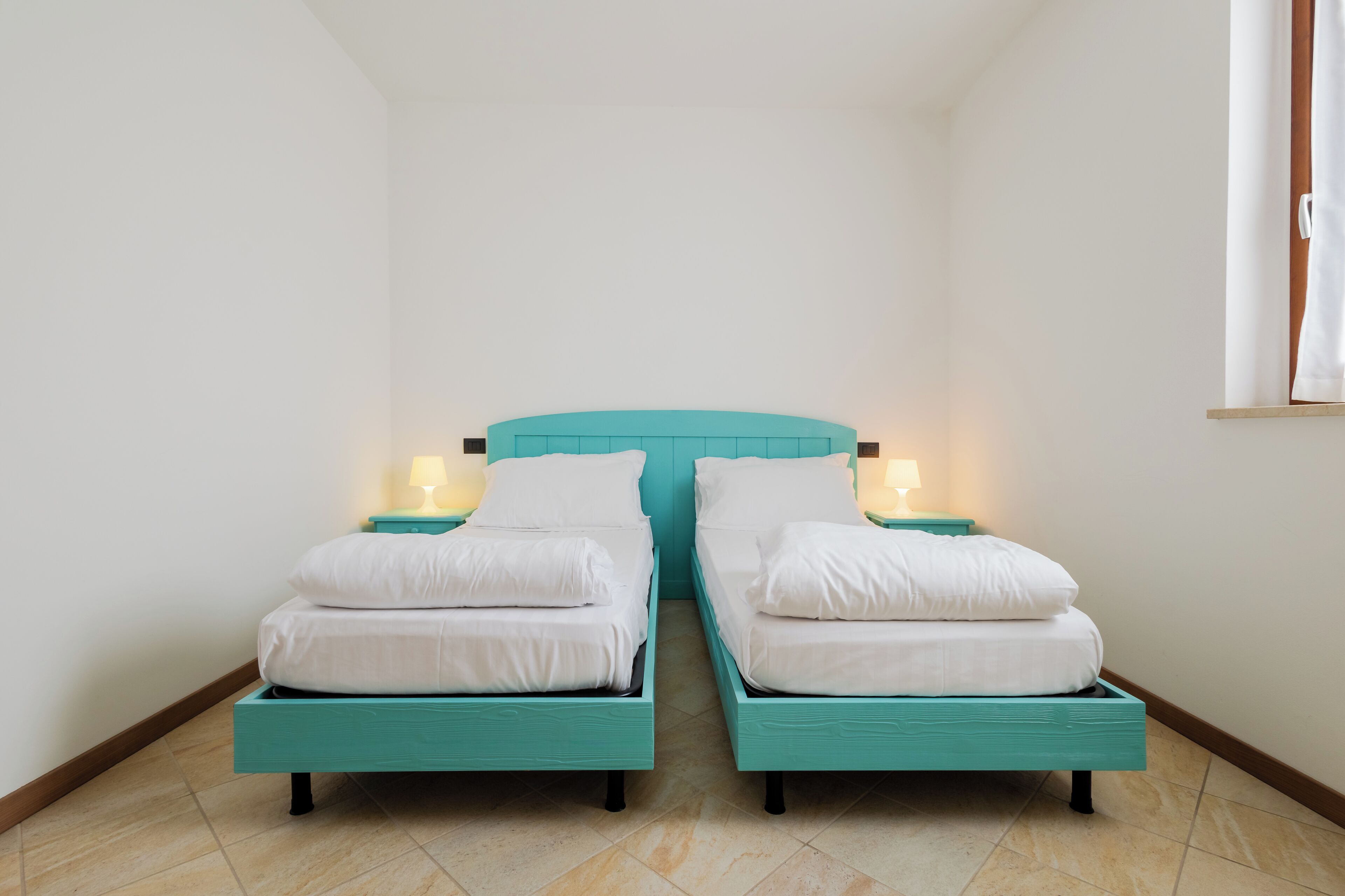 Ein Zimmer mit zwei Betten, jedes mit einem türkisblauen Kopfteil und weißer Bettwäsche. An beiden Seiten der Betten stehen Nachttischlampen.