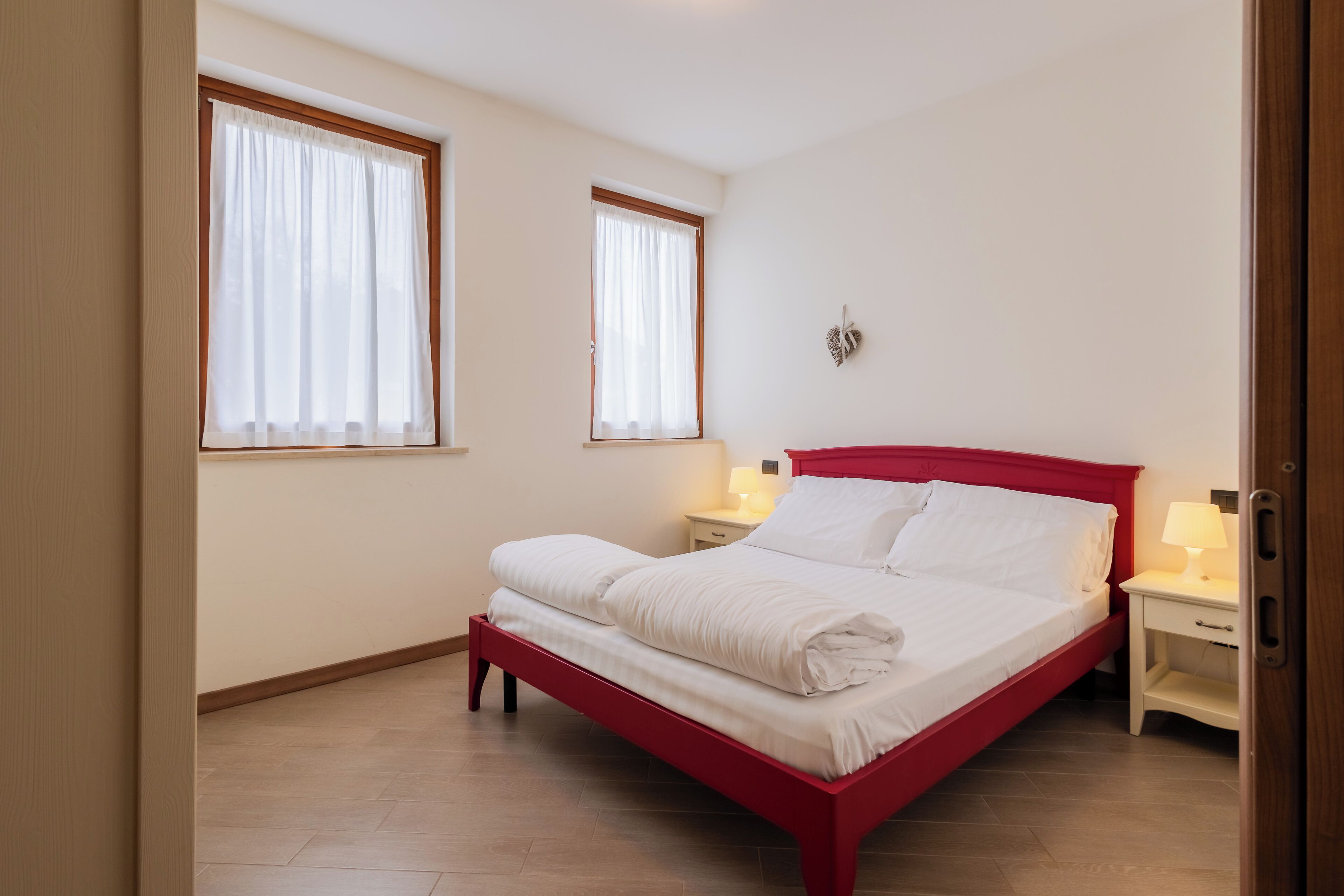 A bedroom with a red bed, two bedside tables with lamps, and two windows with curtains.