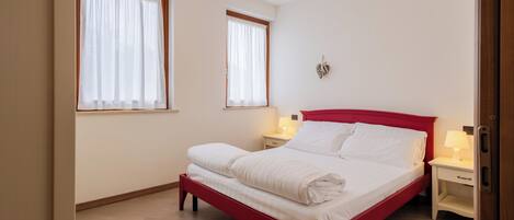 A bedroom with a red bed, two bedside tables with lamps, and two windows with curtains.