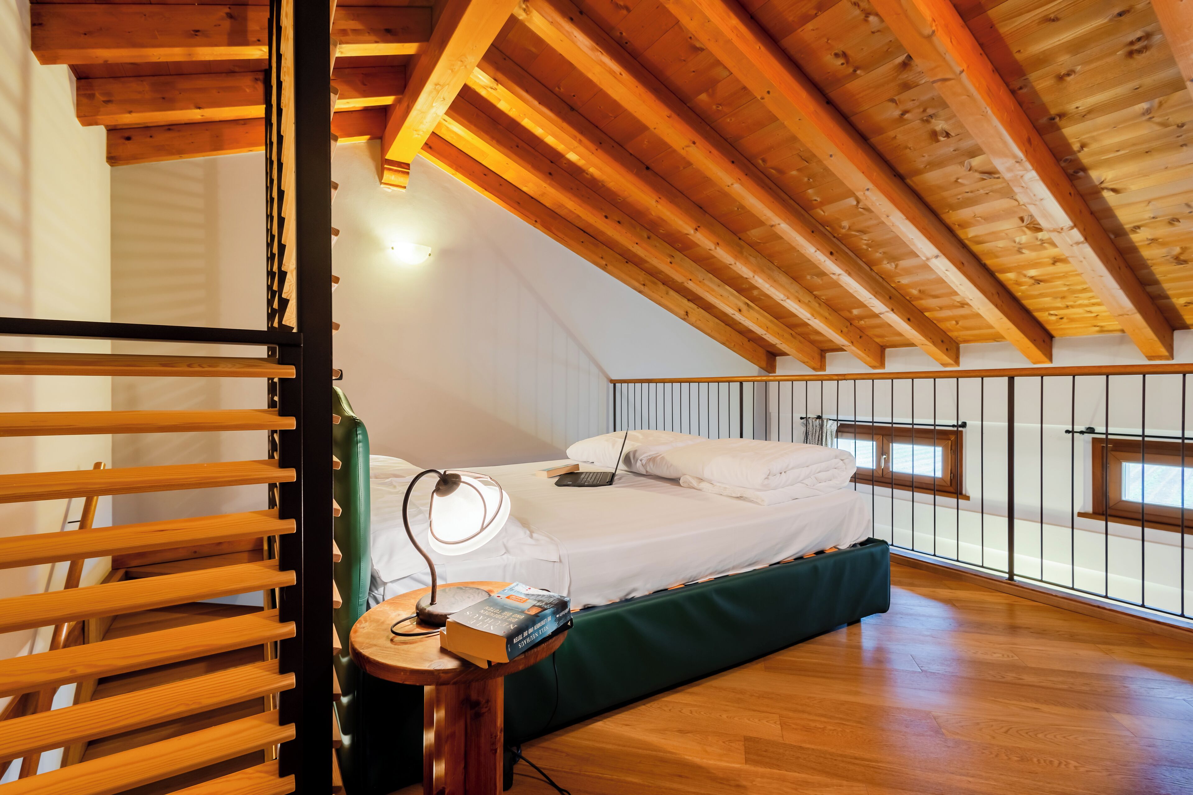 A bedroom with a wooden ceiling, a bed with white bedding, a bedside table with a lamp, and a green bench.