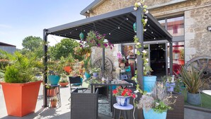 Un patio al aire libre con una pérgola, plantas en macetas, mobiliario de mimbre y un edificio de piedra al fondo.