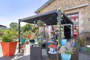 Un patio al aire libre con una pérgola, plantas en macetas, mobiliario de mimbre y un edificio de piedra al fondo.