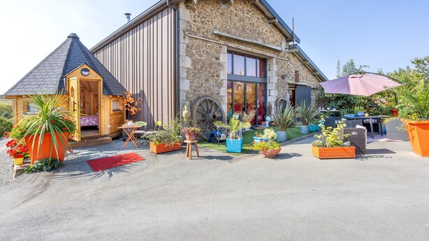 Un edificio de piedra con un anexo de madera, mesas en la terraza y macetas con plantas.