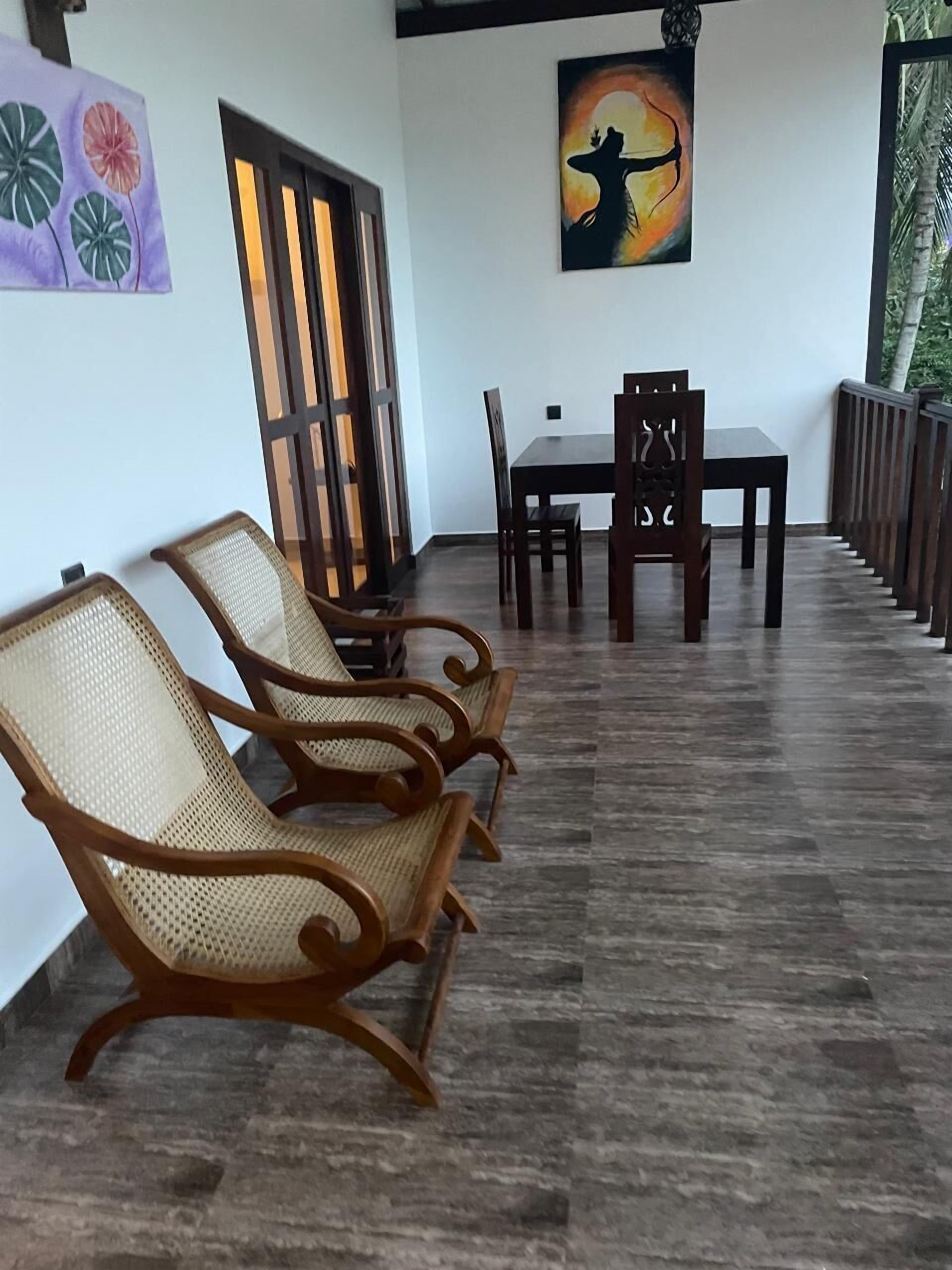 A seating area with wooden chairs and a table, a painting on the wall, and a view of greenery outside.