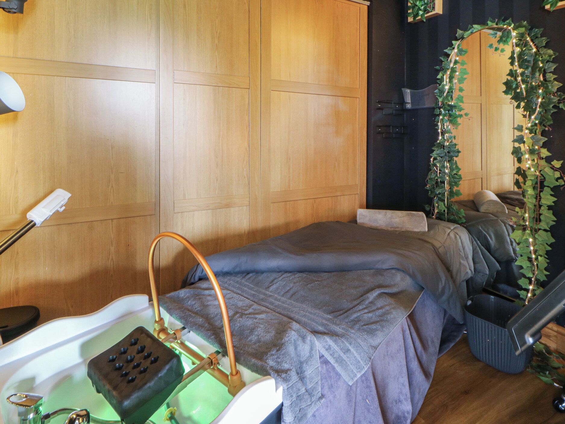 A room with a bed, a small sink with a handheld showerhead, a chair, and a plant.