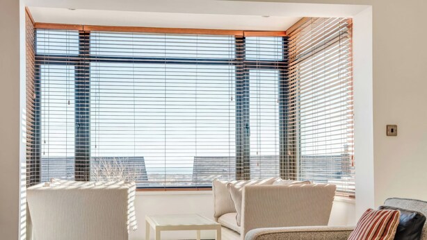 A modern living room with a sofa, armchair, and a glass coffee table. Large windows with blinds provide a view of the cityscape. - Bellevue (Newtownabbey)