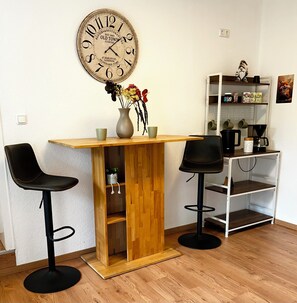 A small kitchen area with a wooden bar counter, two black bar stools, a large wall clock, and a white shelf with kitchen items. - FEWO Unterm Stein - Hänsel & Gretel - 2 sleep. (Alheim)