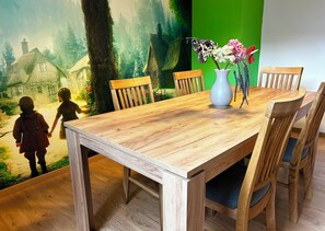 A dining table with wooden chairs, a vase with flowers, and a mural of a forest scene. - FEWO Unterm Stein - Hänsel & Gretel - 2 sleep. (Alheim)