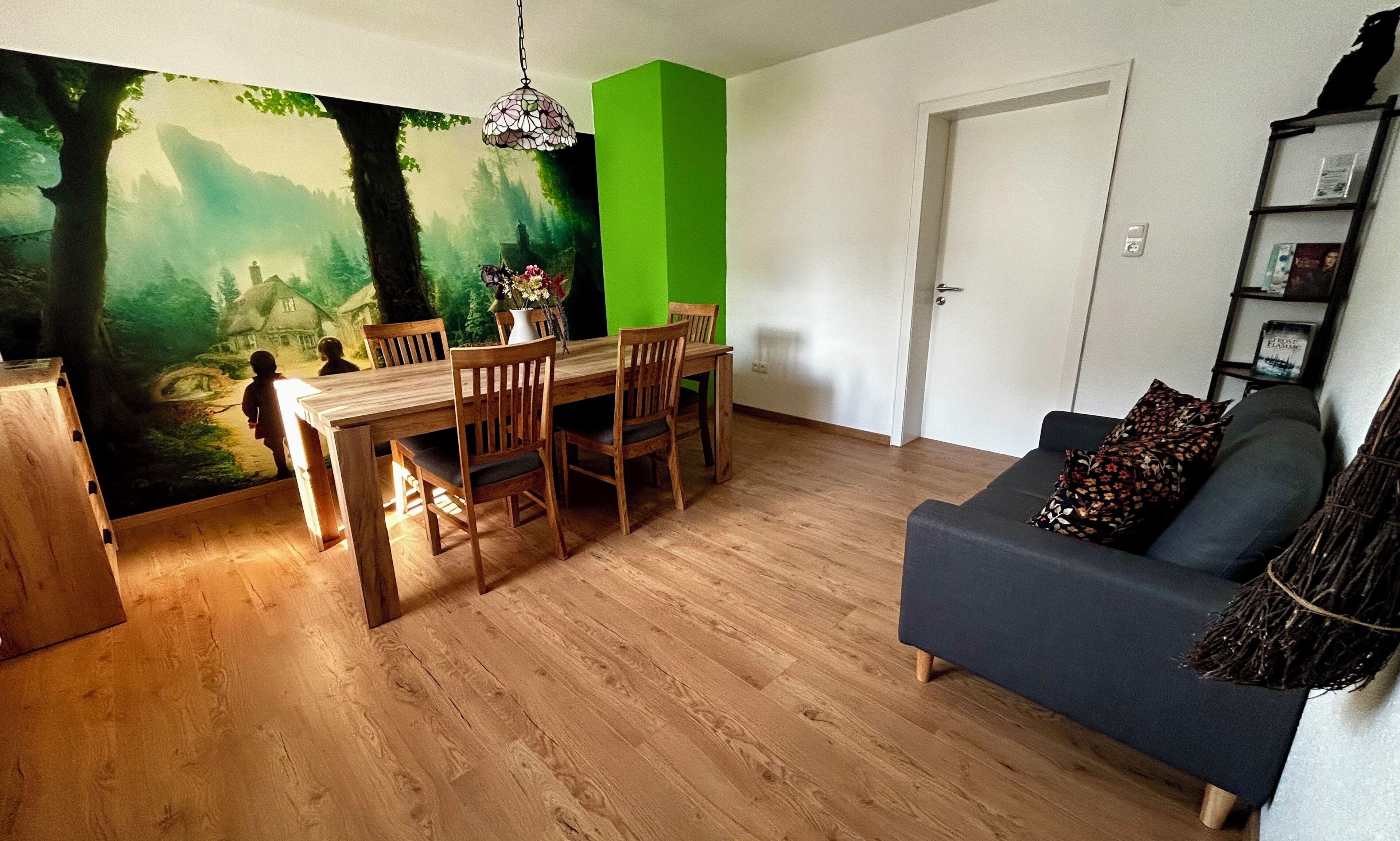 A dining area with a wooden table and chairs, a green accent wall, and a forest mural.