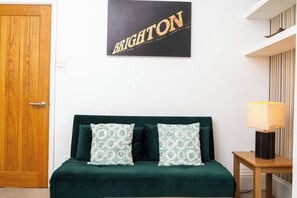 Una habitación con puerta de madera, un sofá verde oscuro con cojines estampados, un cartel en la pared, una mesita con una lámpara y una cortina a rayas.