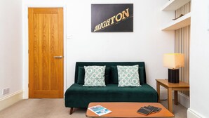 Una habitación con puerta de madera, un sofá verde con cojines estampados, una mesita con una revista y un teléfono, y un cartel en la pared que dice 'BRIGHTON'.