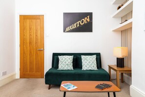 Ein Zimmer mit Holztür, einem grünen Sofa mit gemusterten Kissen, einem kleinen Tisch mit einer Zeitschrift und einem Telefon sowie einem an der Wand befestigten Schild mit der Aufschrift 'BRIGHTON'.