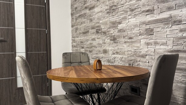 A round wooden table with a central wooden clock, surrounded by four grey upholstered chairs with black wireframe legs. The setting includes a grey brick wall and a wooden door in the background. - Revert Residence - Nespresso, Netflix and Premium Amenities Included (Brașov)