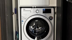 A compact laundry room with a front-loading washing machine, a small shelf above it, and a door to the left. - Revert Residence - Nespresso, Netflix and Premium Amenities Included (Brașov)