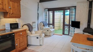 A compact kitchen with a microwave, oven, and wooden cabinets. There is a seating area with a sofa and a table, and a TV mounted on the wall. - Apple Cottage (Bridport)