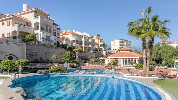 A swimming pool with clear blue water, surrounded by a landscaped area with palm trees and a building with balconies. - Vacation Apartment 'Golf Del Sur Oasis' with Shared Pool, Private Terrace & Wi-Fi (Santa Cruz de Tenerife)
