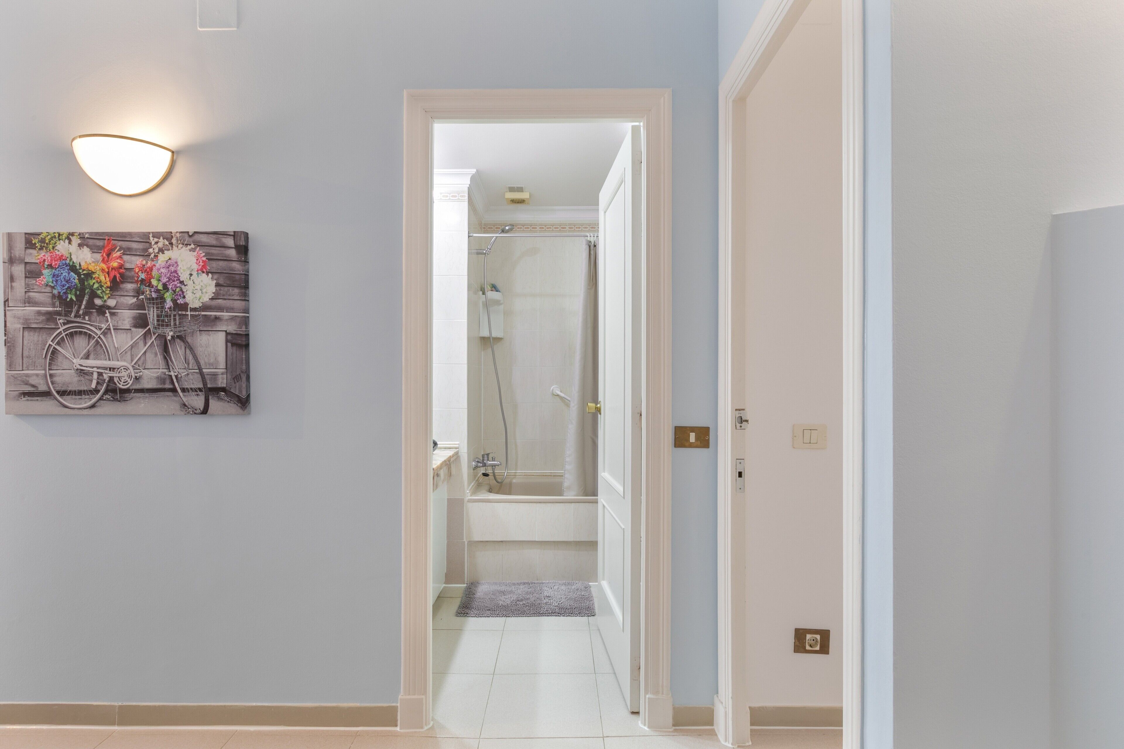 Un pasillo que conduce a un baño con ducha, un accesorio de iluminación y un cuadro de bicicleta colgado en la pared.