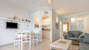 A living room with a sofa, a dining table with chairs, a TV mounted on the wall, and a kitchen area with cabinets and a sink. - Vacation Apartment 'Golf Del Sur Oasis' with Shared Pool, Private Terrace & Wi-Fi (Santa Cruz de Tenerife)