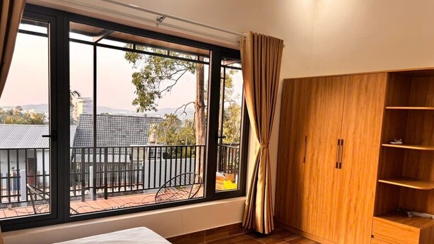 Een slaapkamer met een groot raam, een bed, een houten garderobe en een balkon met uitzicht op gebouwen en bomen.