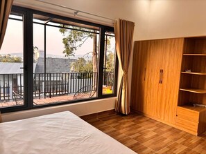 Une chambre avec une grande fenêtre, un lit, une armoire en bois et un balcon offrant une vue sur les immeubles et les arbres.